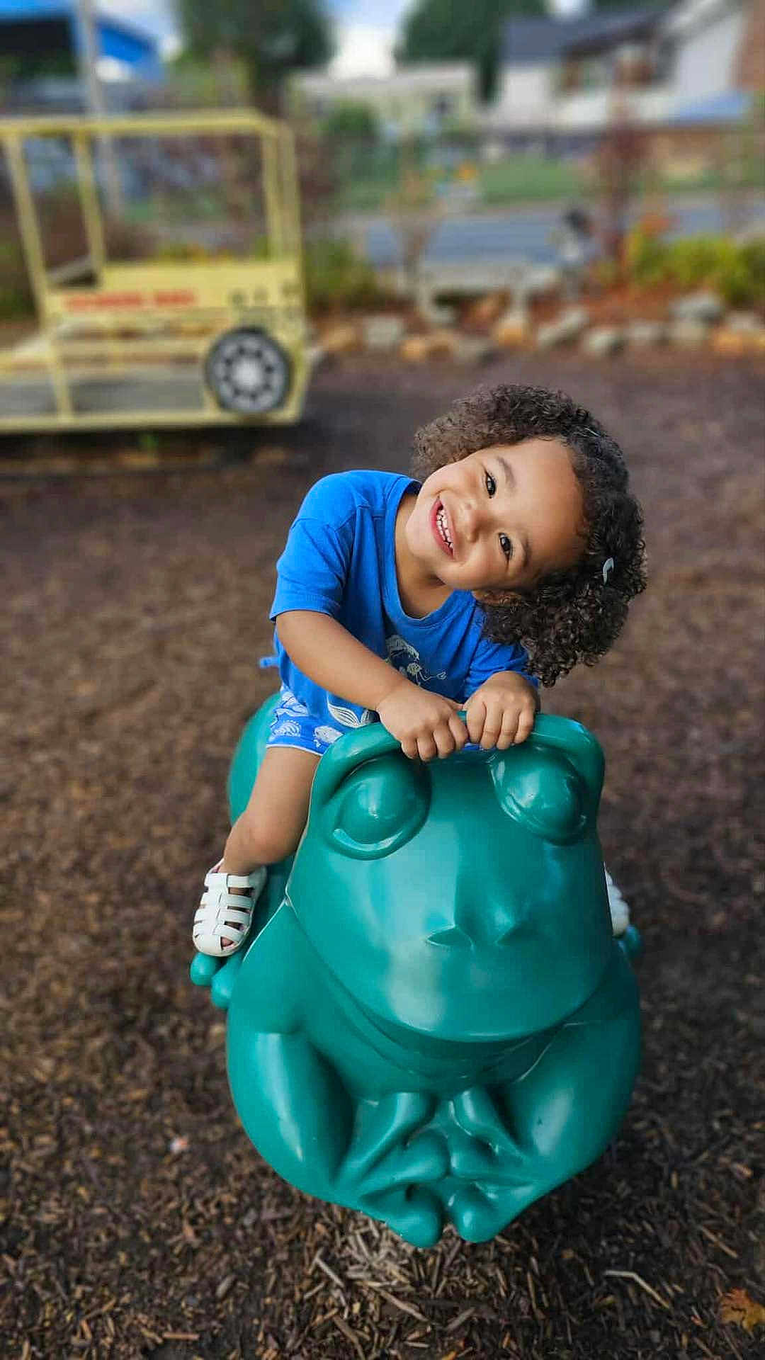 Samaria is registered to the contest to win money with this photo: child, curly_hair, smiling, playground, green_frog, play_toy, blue_shirt, white_shoes, outdoor, park, happy, play, fun, kid, summer, daylight, leisure, recreation, young, person
