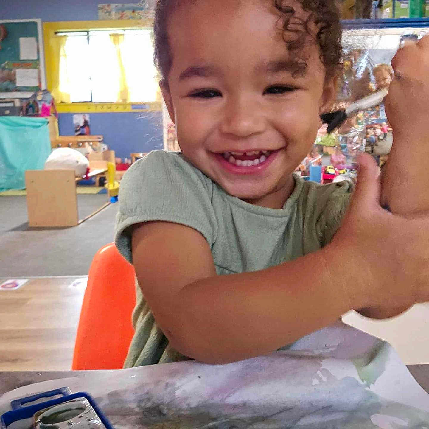 Samaria is registered to the contest to win money with this photo: art_supplies, child, classroom, creative, curly_hair, drawing, expression, happy, indoor, messy, orange_chair, paintbrush, painting, person, playful, smiling, table, toddler, watercolors, young_child