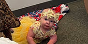 Taylor is registered to the contest to win money with this photo: baby, blanket, carpet, child, clothing, crawling, curious, cute, electrical_outlet, floor, headband, indoor, infant, looking_up, patterned_chair, person, toddler, tutu, wall, yellow