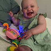 Taylor is registered to the contest to win money with this photo: baby, bed, blanket, child, colorful, cute, foot, green_dress, hand, happy, home, indoor, infant, laughing, person, pillow, playing, smiling, soft_focus, toy