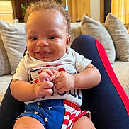 Ashton is registered to the contest to win money with this photo: baby, child, smiling, sitting, indoor, clothing, shorts, tshirt, couch, cushions, person, happy, face, hands, legs, home, living_room, curtains, window, lamp