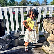 Rylee joined the competition — help win amazing prizes! child, girl, smiling, posing, yellow_cardigan, floral_dress, hair_clips, outdoor, porch, wooden_floor, flower_pots, white_fence, sunlight, shadow, greenery, trees, blue_sky, casual_clothing, happy, playful