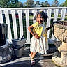 child, girl, smiling, posing, yellow_cardigan, floral_dress, hair_clips, outdoor, porch, wooden_floor, flower_pots, white_fence, sunlight, shadow, greenery, trees, blue_sky, casual_clothing, happy, playful