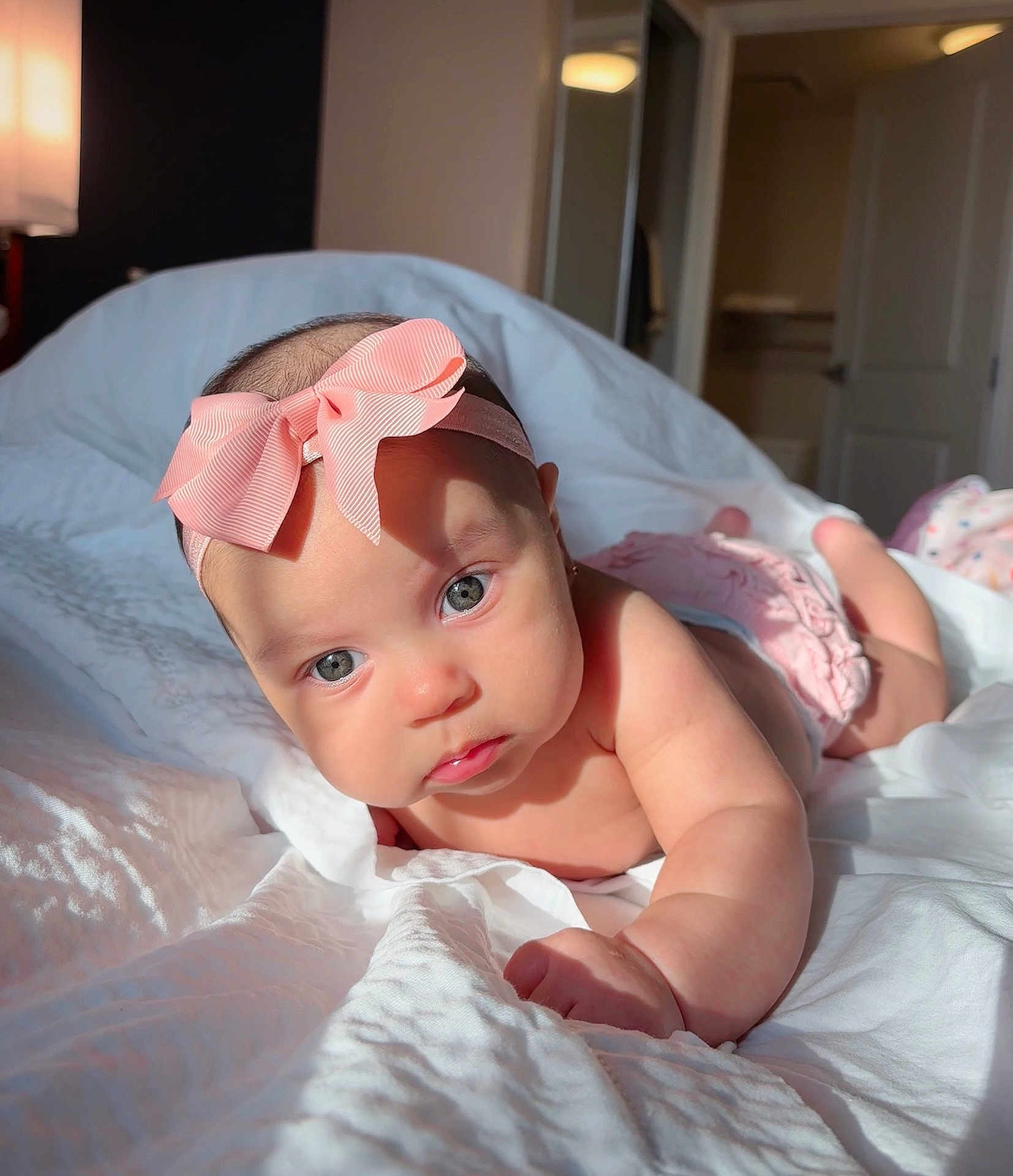 Kensie is registered to the contest to win money with this photo: baby, infant, pink_bow, headband, bed, white_bedspread, sunlight, indoor, portrait, cute, child, skin, face, eyes, expression, room, door, soft_light, laying_down, adorable