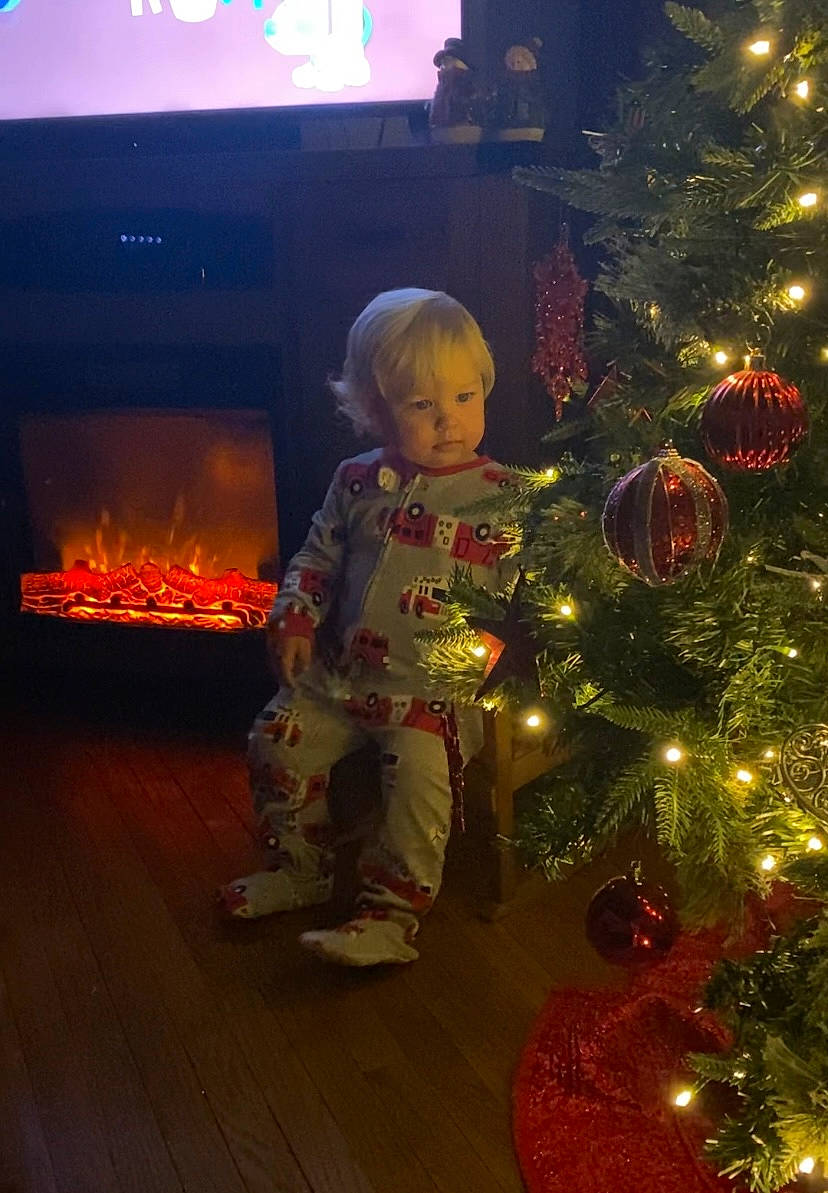 Henry is registered to the contest to win money with this photo: christmas_decoration, christmas_ornament, christmas_tree, conifer, event, evergreen, fun, gas, holiday, holiday_ornament, lap, light, ornament, person, plant, room, standing, toddler, tradition, winter