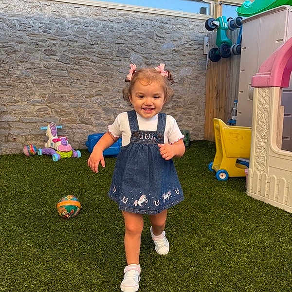 Lenni Kate is registered to the contest to win money with this photo: toddler, child, girl, running, playground, toy, playhouse, grass, artificial_turf, outdoor, smiling, happy, shoes, dress, hair_bows, fence, wall, shade, daylight, recreation