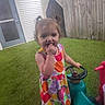 Lenni Kate joined the competition — help win amazing prizes! child, toddler, girl, pigtails, colorful_dress, fruit_pattern, artificial_grass, ride_on_toy, steering_wheel, fence, backyard, door, curious, standing, outdoor, play, hand_in_mouth, young_child, casual, daylight
