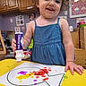 toddler, child, painting, artwork, blue_dress, smiling, yellow_table, indoor, toy, colorful, creative, playroom, face, person, hand, paper, bottle, happy, expression, young_child