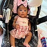 baby, black_hair, car_seat, cherry_print, child, curious_expression, cute, fabric_texture, footwear_absent, head_support, indoor, infant, patterned_fabric, pink_bow, pink_dress, playful, portrait, seat_belt, toy, white_toy