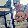 baby, infant, child, glasses, onesie, elephant_print, hand, furniture, window, blinds, curious, indoor, person, cute, toddler, red_dress, shelf, toy, home, portrait