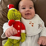 Jalil participe au concours pour gagner de l'argent avec cette photo : baby, infant, plush_toy, grinch, red_hat, white_bib, smile, brown_pillow, cozy, cute, child, soft_toy, holiday_theme, red_clothing, white_outfit, hand, face, portrait, lying_down, indoor