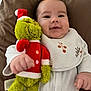 baby, infant, plush_toy, grinch, red_hat, white_bib, smile, brown_pillow, cozy, cute, child, soft_toy, holiday_theme, red_clothing, white_outfit, hand, face, portrait, lying_down, indoor