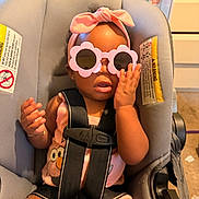 Sevyn is registered to the contest to win money with this photo: baby, child, car_seat, sunglasses, headband, pink, straps, indoor, person, cute, expression, clothing, seat, hand, face, infant, accessory, room, furniture, toy