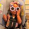 baby, child, car_seat, sunglasses, headband, pink, straps, indoor, person, cute, expression, clothing, seat, hand, face, infant, accessory, room, furniture, toy