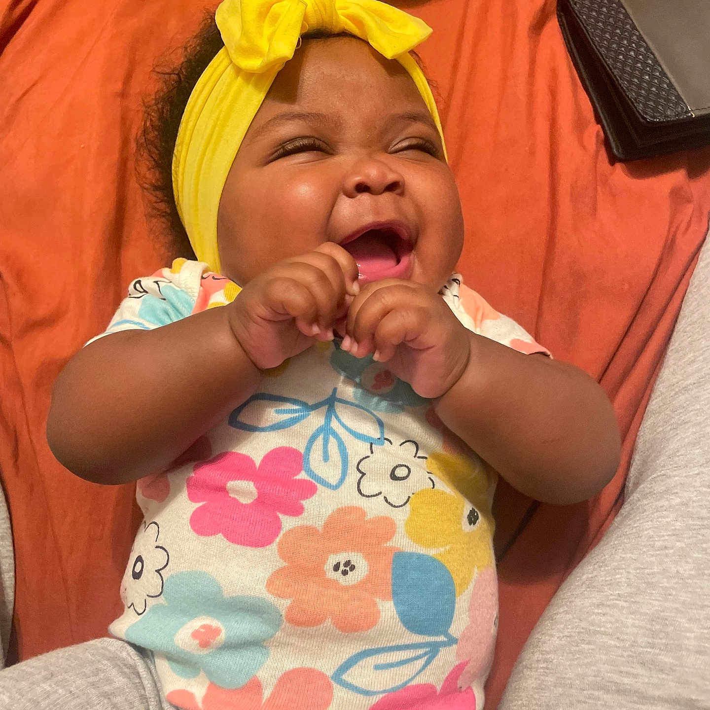 Umharia is registered to the contest to win money with this photo: baby, child, smiling, laughing, yellow_bow, headband, floral_shirt, colorful_clothing, gray_pants, orange_blanket, wallet, patterned_objects, happy, cute, infant, portrait, indoors, hands, joy, person