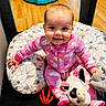 baby, child, smiling, pink_onesie, plush_toy, cow_toy, stuffed_animal, cushion, floral_pattern, floor, wooden_floor, indoor, happy, cute, infant, sitting, playpen, toy, person, face