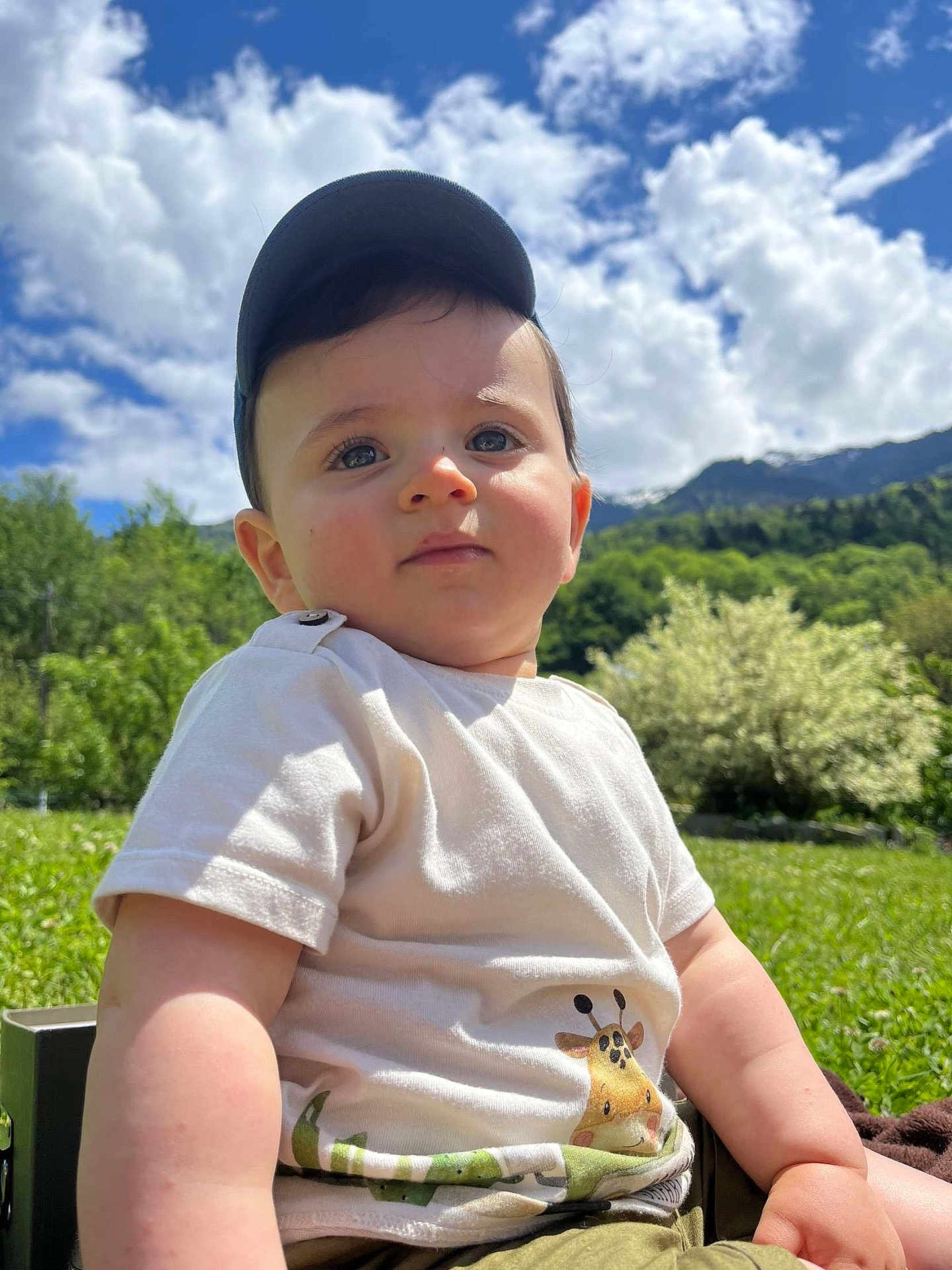 Karl participe au concours pour gagner de l'argent avec cette photo : baby, child, cap, tshirt, giraffe_print, outdoor, nature, grass, mountain, sky, clouds, sunny, greenery, portrait, cute, sitting, daylight, person, face, young