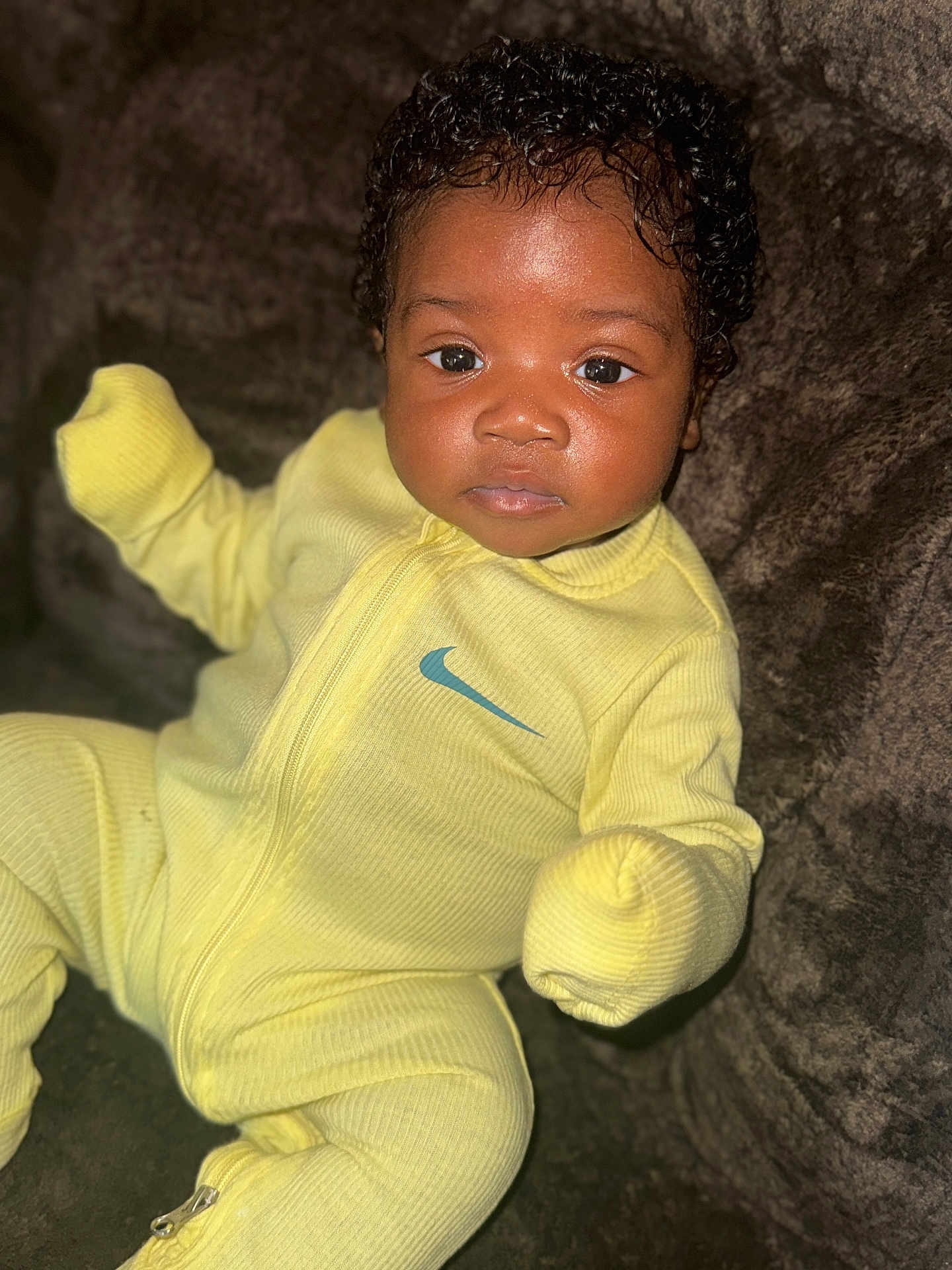 Jaikhyius joined the competition — help win amazing prizes! baby, child, infant, yellow_onesie, nike_logo, curly_hair, sitting, soft_texture, brown_background, portrait, face, eyes, cute, clothing, warm, indoors, young_child, looking_at_camera, cozy, comfortable