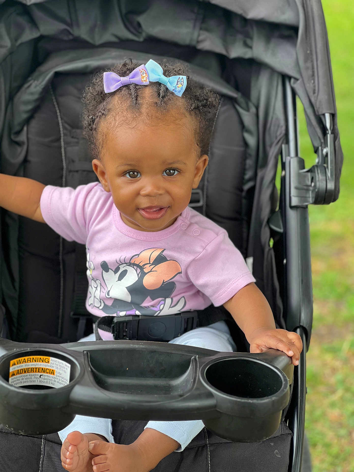 Seven is registered to the contest to win money with this photo: toddler, child, stroller, smile, bow, hair, clothing, shirt, minnie_mouse, baby, outdoor, seatbelt, foot, hand, person, cute, happy, portrait, playful, grass