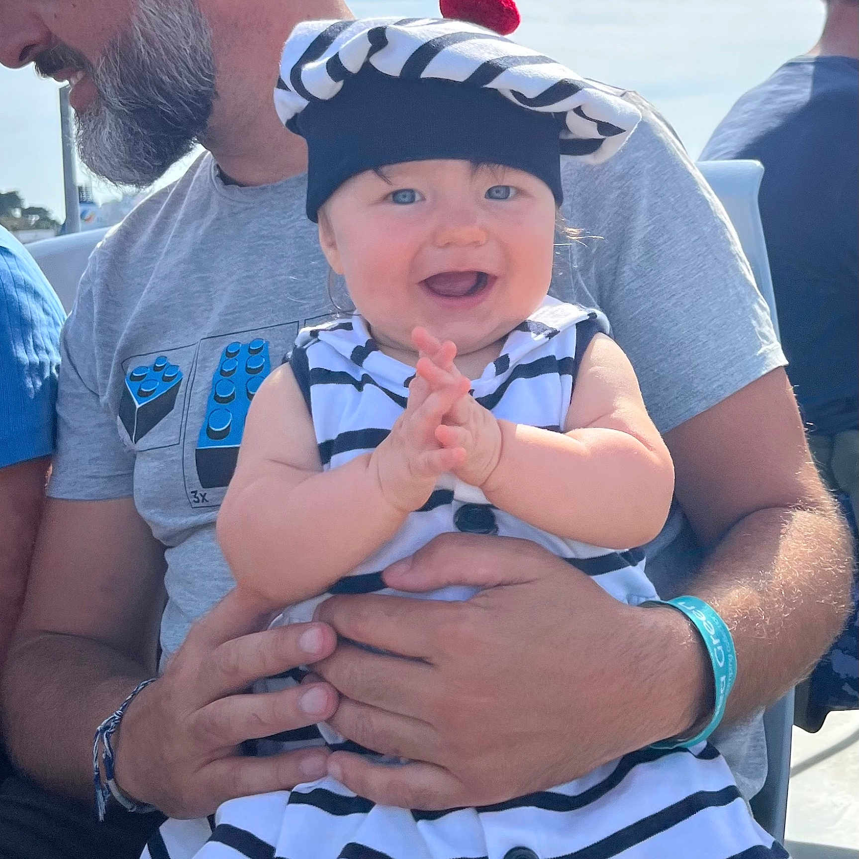 Mélyne participe au concours pour gagner de l'argent avec cette photo : baby, beard, beret, blue_eyes, casual_wear, child, clapping, daylight, facial_expression, hands, happy, holding, man, outdoor, person, red_pom_pom, seated, short_sleeves, smiling, striped_clothing