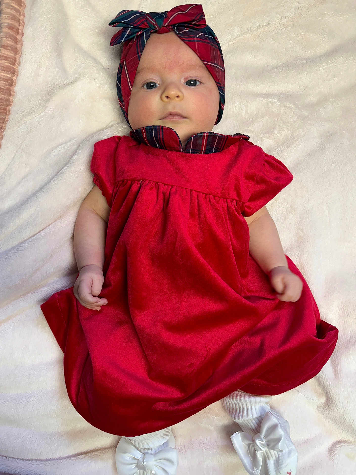 Anahi is registered to the contest to win money with this photo: baby, infant, red_dress, headband, plaid, white_socks, bow, blanket, soft_texture, lying_down, portrait, cute, child, young, face, hands, footwear, cozy, indoor, fashion