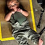 baby, child, green_clothing, overalls, wrench, tool, floor, mat, yellow_border, concrete, barefoot, face, hands, playful, lying_down, workshop, mechanic, cute, indoor, person