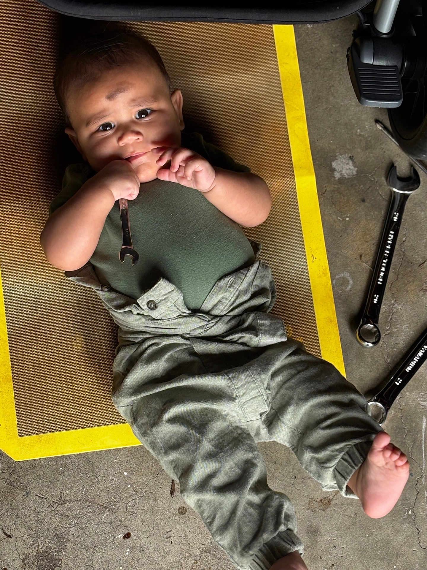 Henry joined the competition — help win amazing prizes! baby, child, green_clothing, overalls, wrench, tool, floor, mat, yellow_border, concrete, barefoot, face, hands, playful, lying_down, workshop, mechanic, cute, indoor, person