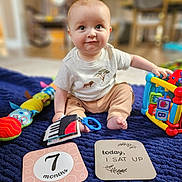 Hogan is registered to the contest to win money with this photo: baby, child, infant, milestone_cards, toy, colorful_toy, blanket, sitting, smiling, indoor, cute, playtime, person, young_child, milestone, home, floor, happy, portrait, clothing