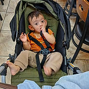 Muzammil is registered to the contest to win money with this photo: baby, barefoot, beige_pants, black_chair, cafe, casual_clothing, chair, child, curious, floor, furniture, indoor, orange_shirt, person, portrait, seatbelt, stroller, tiles, toddler, wooden_chair