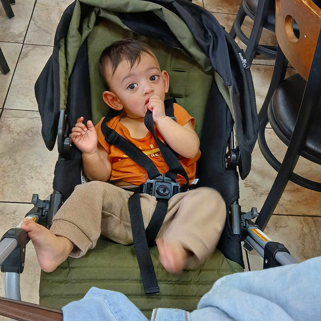 Muzammil is registered to the contest to win money with this photo: baby, barefoot, beige_pants, black_chair, cafe, casual_clothing, chair, child, curious, floor, furniture, indoor, orange_shirt, person, portrait, seatbelt, stroller, tiles, toddler, wooden_chair