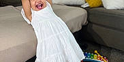 Gleiany is registered to the contest to win money with this photo: toddler, child, white_dress, black_sandals, smiling, happy, indoor, living_room, couch, pillows, toys, carpet, playing, person, young_child, joyful, colorful, xylophone, furniture, home