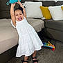 toddler, child, white_dress, black_sandals, smiling, happy, indoor, living_room, couch, pillows, toys, carpet, playing, person, young_child, joyful, colorful, xylophone, furniture, home