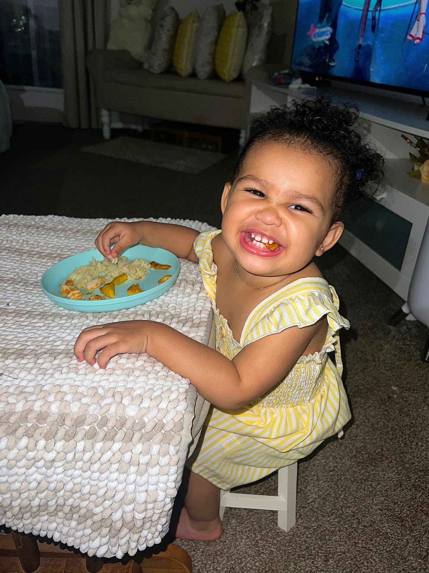 Gleiany joined the competition — help win amazing prizes! toddler, child, smiling, eating, food, plate, table, chair, yellow_dress, curly_hair, living_room, carpet, sofa, pillows, television, happy, indoor, person, candid, meal