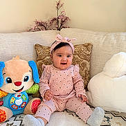 Anaya joined the competition — help win amazing prizes! baby, child, cushion, cute, decor, face, flower, happy, headband, indoor, infant, patterned_rug, person, pink_clothing, plush_toy, sitting, smiling, sock, sofa, toy