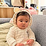 Anaya is registered to the contest to win money with this photo: baby, blanket, child, couch, cozy, cute, ears, face, hand, hood, indoor, infant, kitchen, long_hair, onesie, person, pink_bow, sitting, soft_fabric, window
