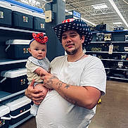 Giovanni is registered to the contest to win money with this photo: baby, man, cowboy_hat, red_hat, store, aisle, coolers, shelves, party_supplies, tattoos, white_tshirt, holding, headband, indoor, shopping, curious, portrait, people, floor, lighting
