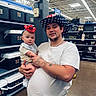 baby, man, cowboy_hat, red_hat, store, aisle, coolers, shelves, party_supplies, tattoos, white_tshirt, holding, headband, indoor, shopping, curious, portrait, people, floor, lighting