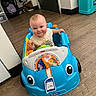 baby, child, toy_car, blue_toy, smiling, indoor, floor, wood_floor, face, person, clothing, play, fun, colorful, alphabet, car_seat, door, cooler, shoes, baby_toy