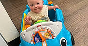 Giovanni is registered to the contest to win money with this photo: baby, toy_car, indoor, floor, wood_floor, child, smiling, blue, plastic_toy, face, person, play, cute, infant, small_car, room, door, cooler_box, clothing, happy