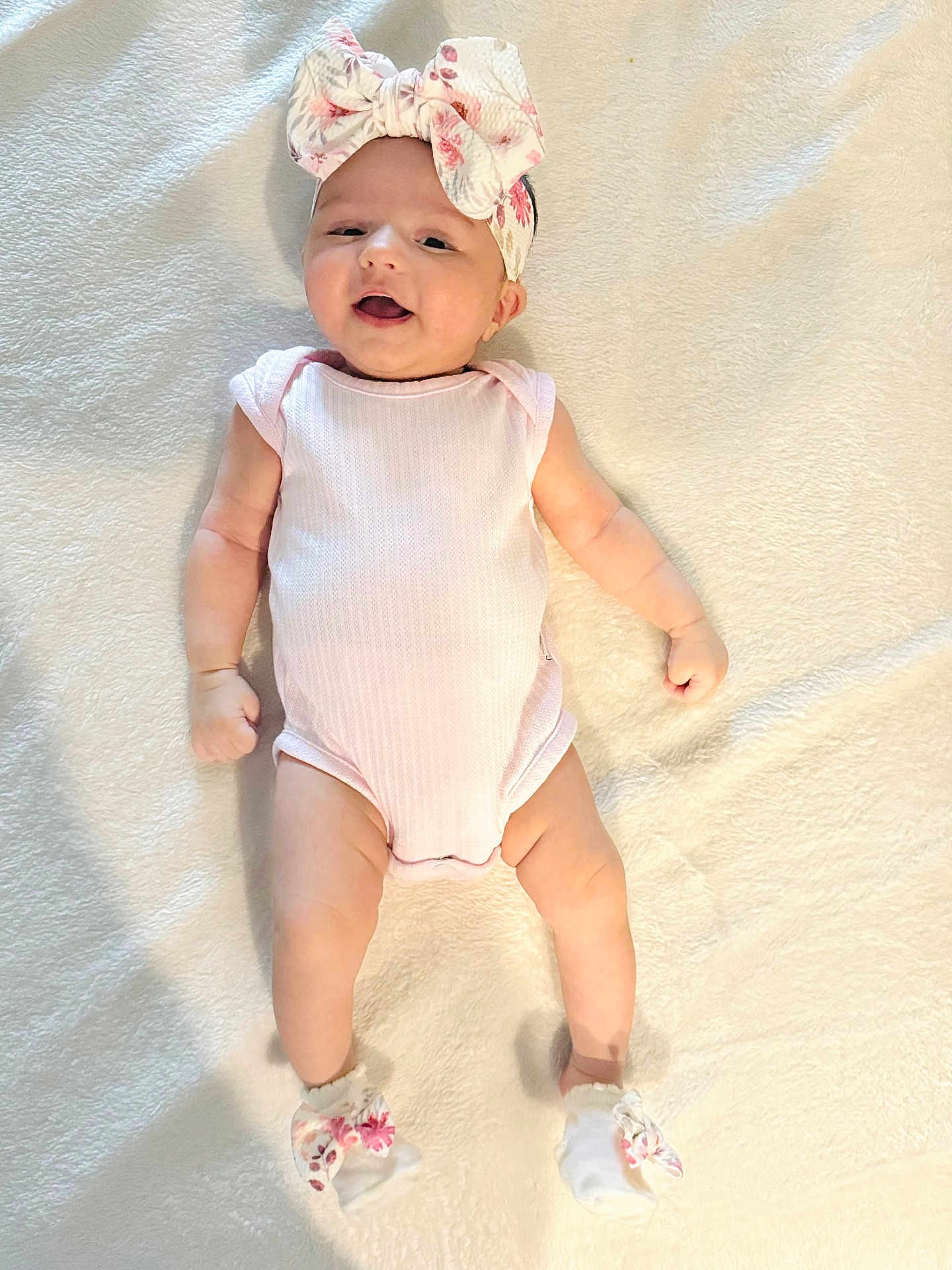 Minha is registered to the contest to win money with this photo: baby, infant, smiling, headband, bow, socks, lying_down, blanket, cute, happy, child, soft_texture, clothing, person, newborn, pink, floral, indoors, portrait, baby_girl