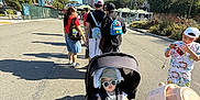 Rayleigh Lucas is registered to the contest to win money with this photo: baby, stroller, child, people, backpack, pajamas, toy, sunglasses, hat, road, outdoor, sunny, shadows, trees, palm_tree, sky, group, walking, casual_clothing, daytime