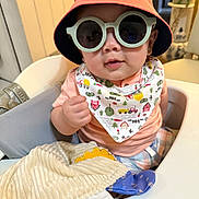 Rayleigh Lucas is registered to the contest to win money with this photo: baby, sunglasses, hat, bib, high_chair, toy, indoor, child, cute, smiling, face, clothing, person, seat, accessory, infant, dinosaur_toy, plush, orange, patterned
