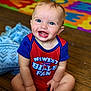 baby, child, smiling, blue_eyes, onesie, red_clothing, blue_clothing, sitting, wooden_floor, blanket, colorful_mat, indoor, happy, cute, portrait, person, infant, playful, young_child, casual