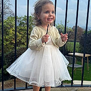Goldie is registered to the contest to win money with this photo: child, toddler, dress, white_dress, golden_shoes, outdoor, balcony, railing, smiling, curly_hair, happy, grass, table, chair, stone_floor, nature, person, fashion, portrait, cute