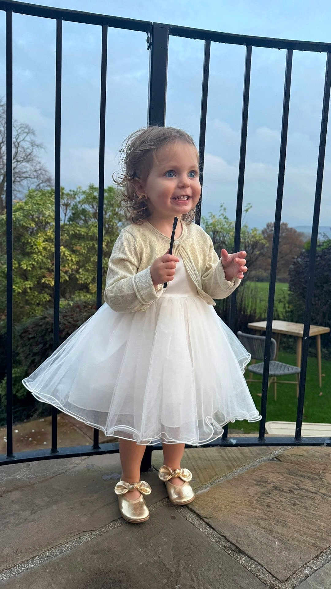 Goldie is registered to the contest to win money with this photo: child, toddler, dress, white_dress, golden_shoes, outdoor, balcony, railing, smiling, curly_hair, happy, grass, table, chair, stone_floor, nature, person, fashion, portrait, cute