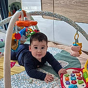 Marcio participe au concours pour gagner de l'argent avec cette photo : baby, child, play_mat, toy, educational_toy, hanging_toys, indoor, floor, wooden_cabinet, window, natural_light, colorful, curious, clothing, smiling, person, infant, crawling, soft_toys, home