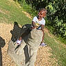 child, toddler, outdoor, statue, elephant, zoo, park, braids, smiling, sitting, riding, sandal, t_shirt, playful, sunlight, shadow, greenery, grass, gravel, portrait