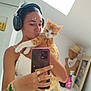 woman, cat, selfie, mirror, headphones, smartphone, orange_cat, white_cat, indoor, casual_clothing, bracelets, rings, mannequin, hat, bandana, skylight, toilet, spray_bottle, mirror_reflection, bedroom