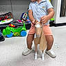 toddler, child, toy_horse, toy, indoor, store, floor, shelf, clothing, shorts, polo_shirt, footwear, curly_hair, play, plastic_toy, looking_away, white_shoes, floor_tiles, retail, shopping_aisle