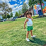 Adam joined the competition — help win amazing prizes! child, toddler, grass, park, outdoor, sunny, clouds, blue_sky, tree, leaves, green_shorts, white_shoes, white_tank_top, curly_hair, mural, colorful, chairs, playground, daytime, person